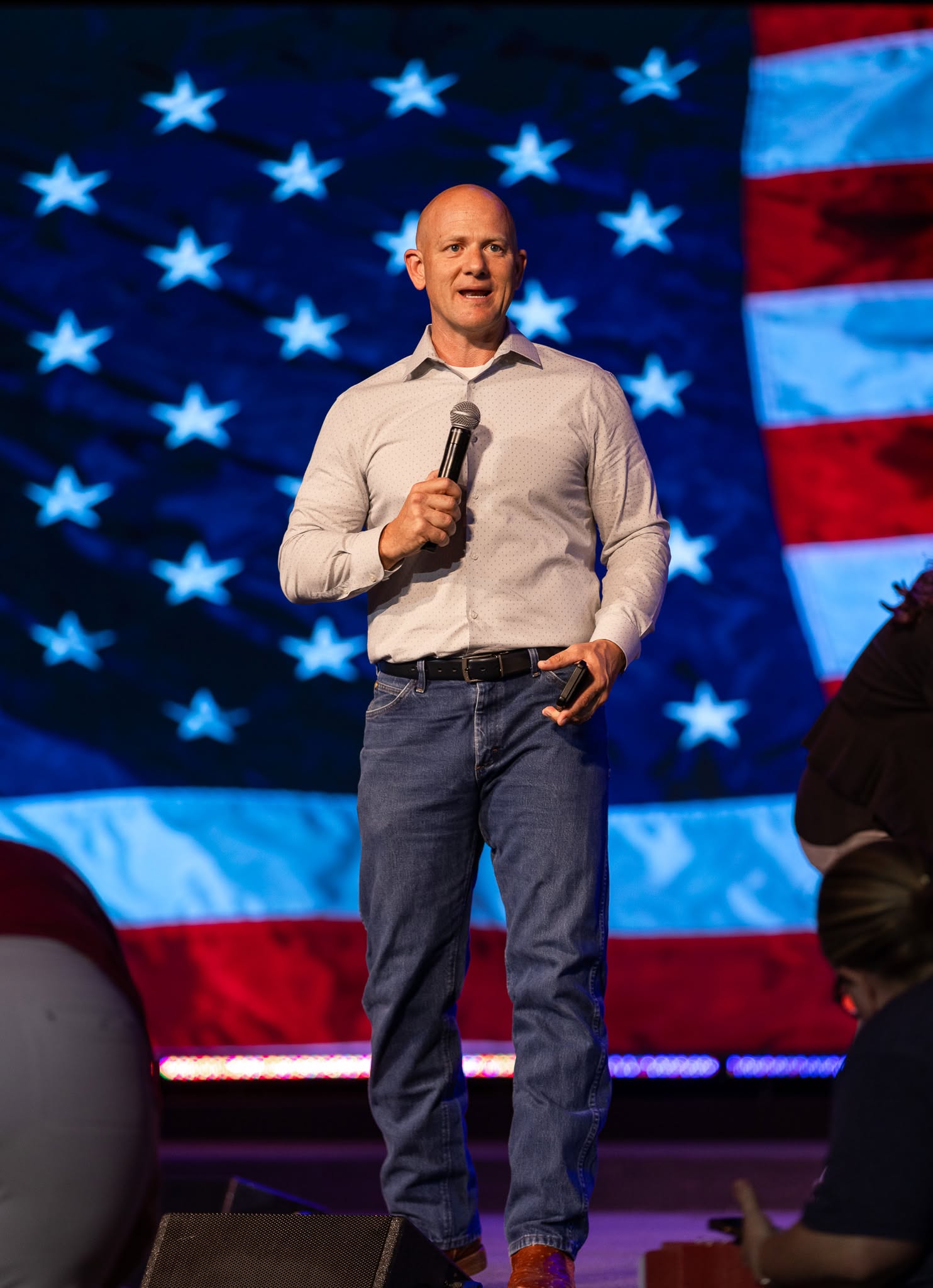 Jake Merrick speaking on stage in front of an American flag